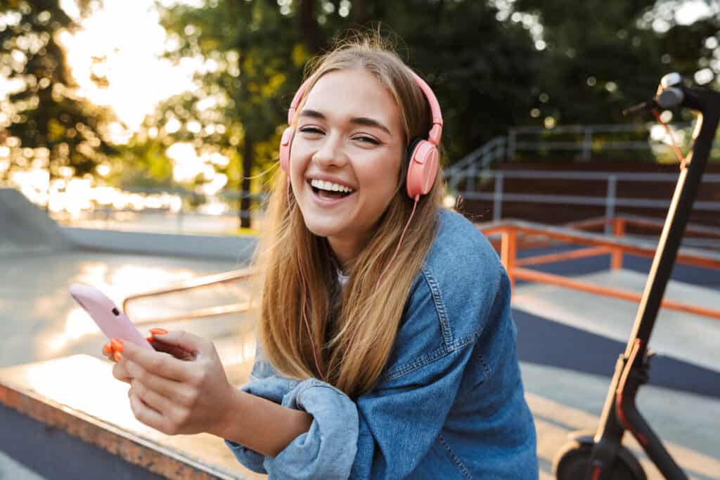 Smiling young woman with headphones using smartphone outdoors near skatepark.